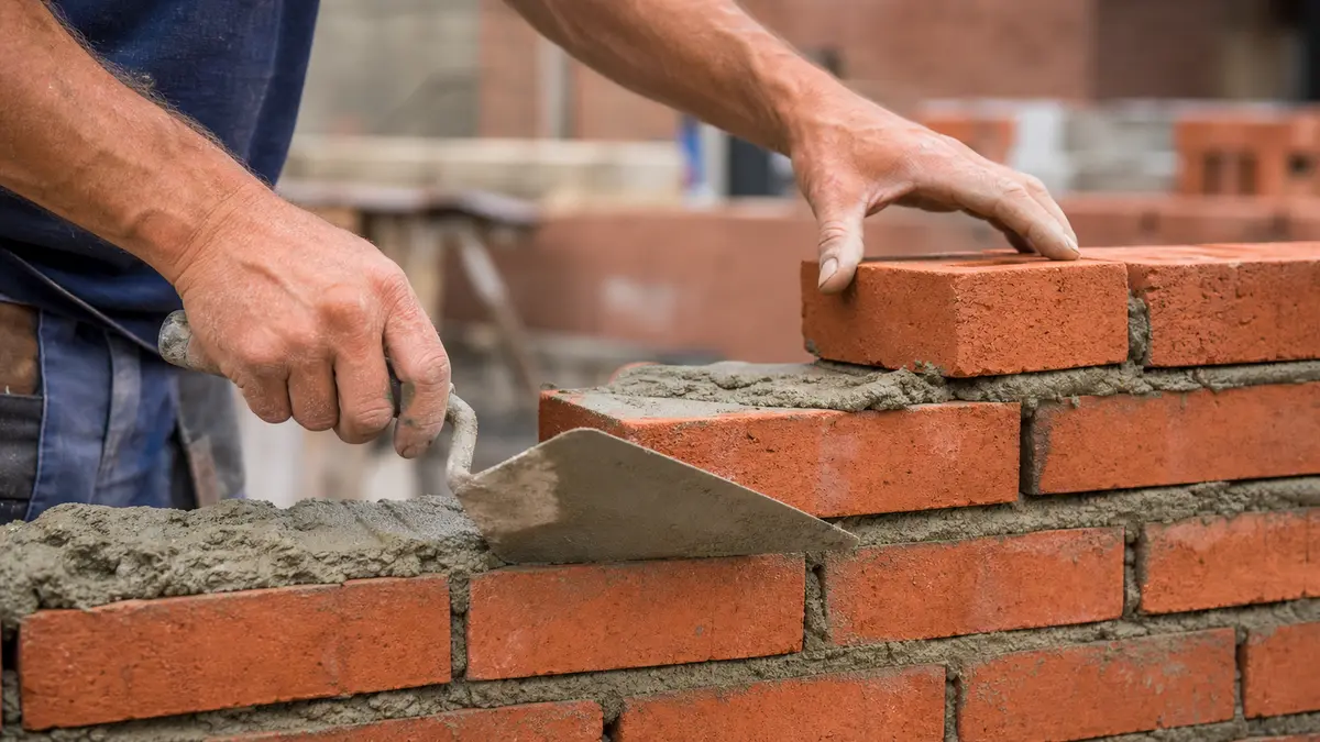 A mason laying modular face bricks in a running bond pattern with a trowel and mortar on a wall